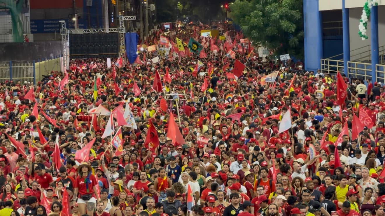 Multidão com camisetas e bandeiras vermelhas ocupa uma avenida durante ato político.