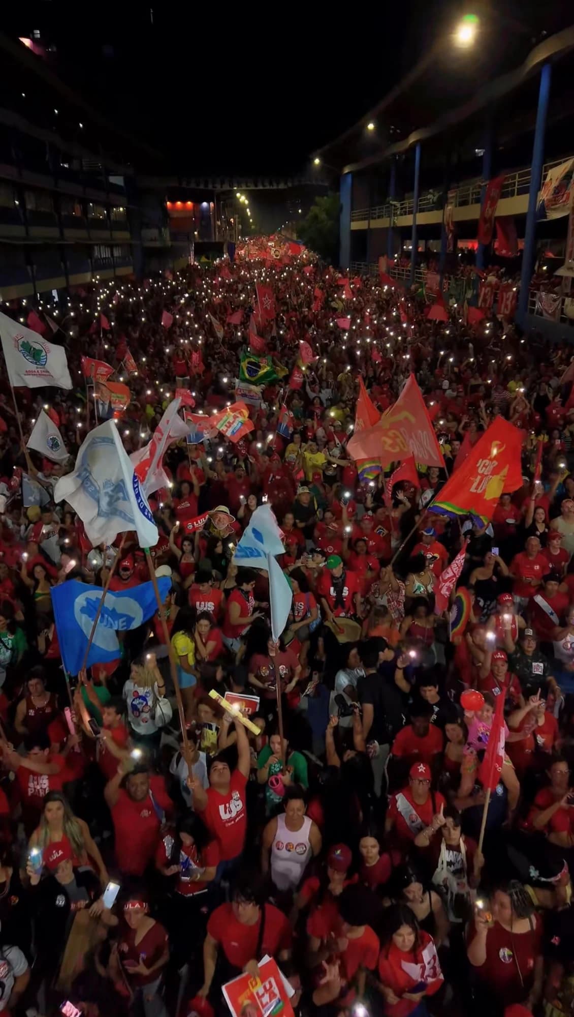 Vista aérea, à noite, de uma multidão com bandeiras vermelhas e luzes de celulares acesas ao longo de uma avenida.